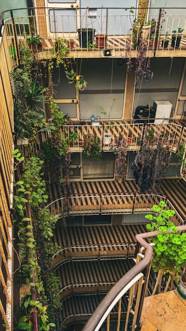 A cozy balcony garden scene with a wooden ledge holding several small terracotta pots containing various green plants, including a succulent and leafy foliage. Behind the pots, a dark glass bottle wit